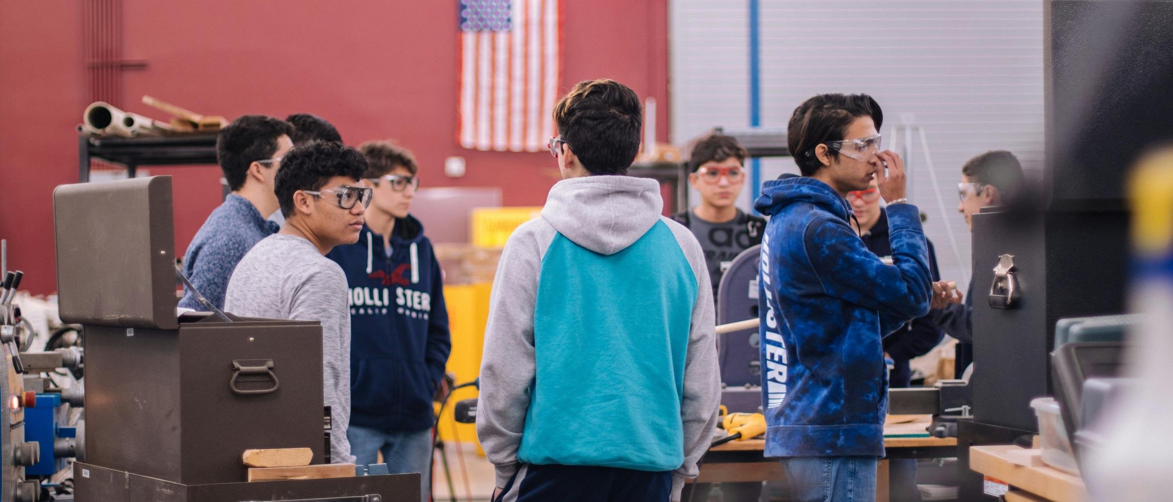 A group of high school students touring a technical shop floor with an American flag on the wall