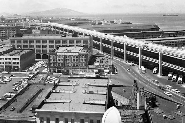 Aerial photograph showing the double-deck Embarcadero Freeway elevated over the working San Francisco waterfront, with finger piers and industrial buildings below. The freeway runs along the city's eastern edge near the Ferry Building, walling off the northern piers from street-level access.